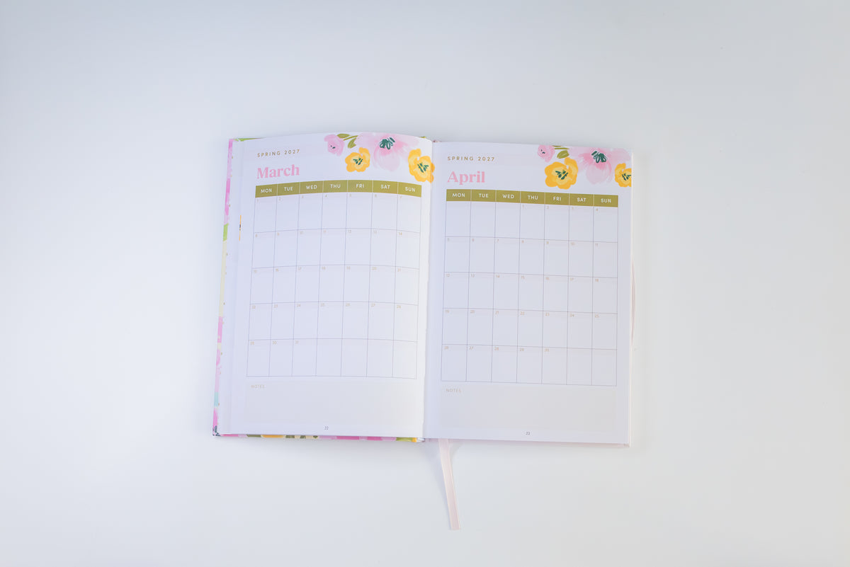 An open planner or agenda showing the monthly grids for March and April, with a pastel floral cover and ribbon bookmark, photographed on a clean white background.