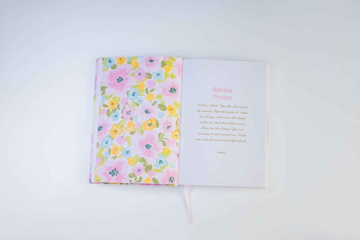 An open journal or prayer book on a clean white background. The left page has a pastel floral cover with pink, yellow, and blue flowers, and the right page is white with a pink title reading 'Spring Prayer' and a short prayer in pink script.