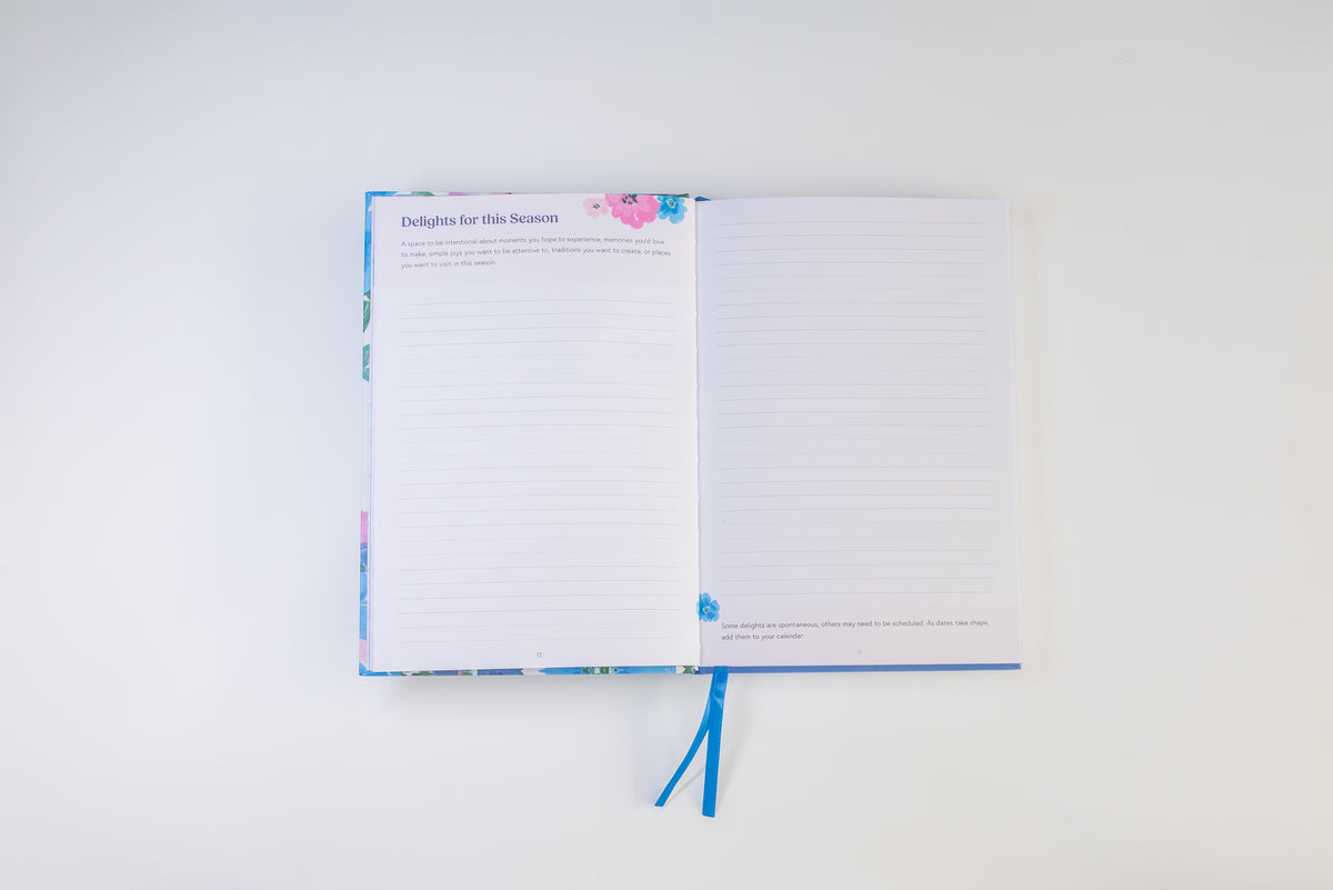 An open lined notebook with a blue ribbon bookmark, photographed from above on a clean white background. The cover and inner pages have a light blue base with small pink and purple floral accents.