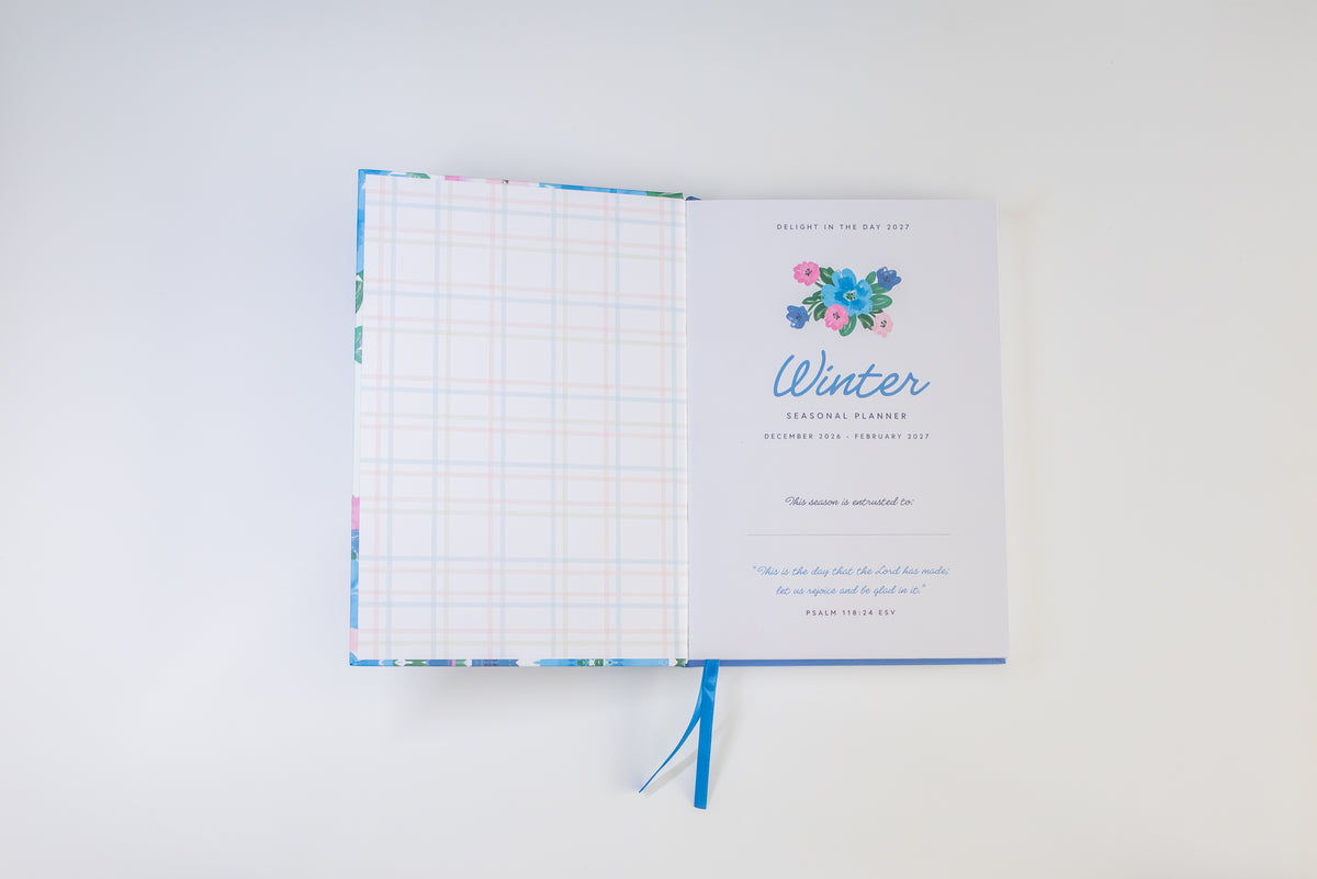 An open winter-themed journal or planner on a clean white background. The left page shows a pastel grid pattern, and the right page features a floral illustration above the word 'Winter' in blue script, with a blue ribbon bookmark hanging below.