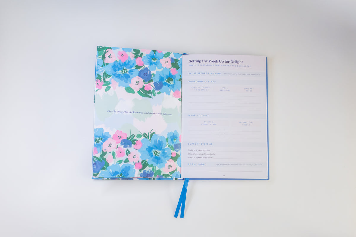 An open planner or journal with a floral cover in blue, pink, and green, showing a printed weekly planning page on the right and a blue ribbon bookmark below, photographed on a clean white background.
