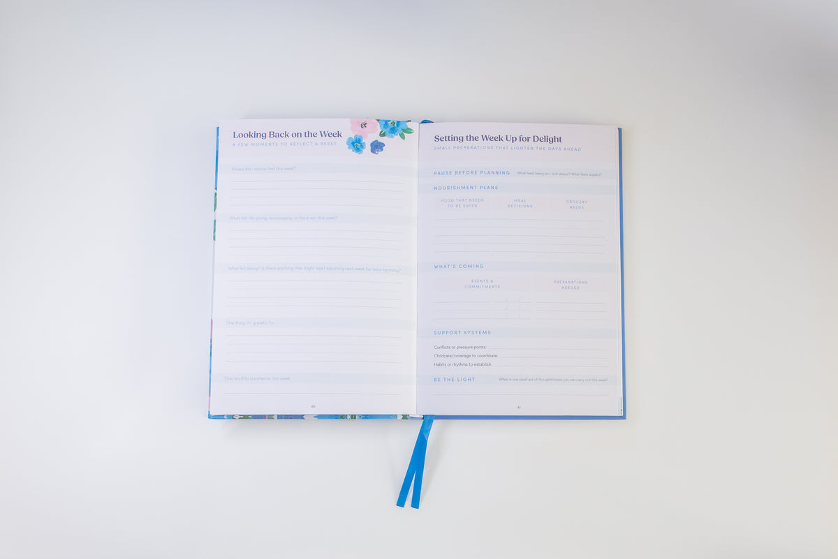 An open planner or notebook with two printed interior pages, shown on a clean white background. The pages are white with light blue text and lines, and the cover has a small floral print with blue ribbon bookmarks hanging from the bottom.