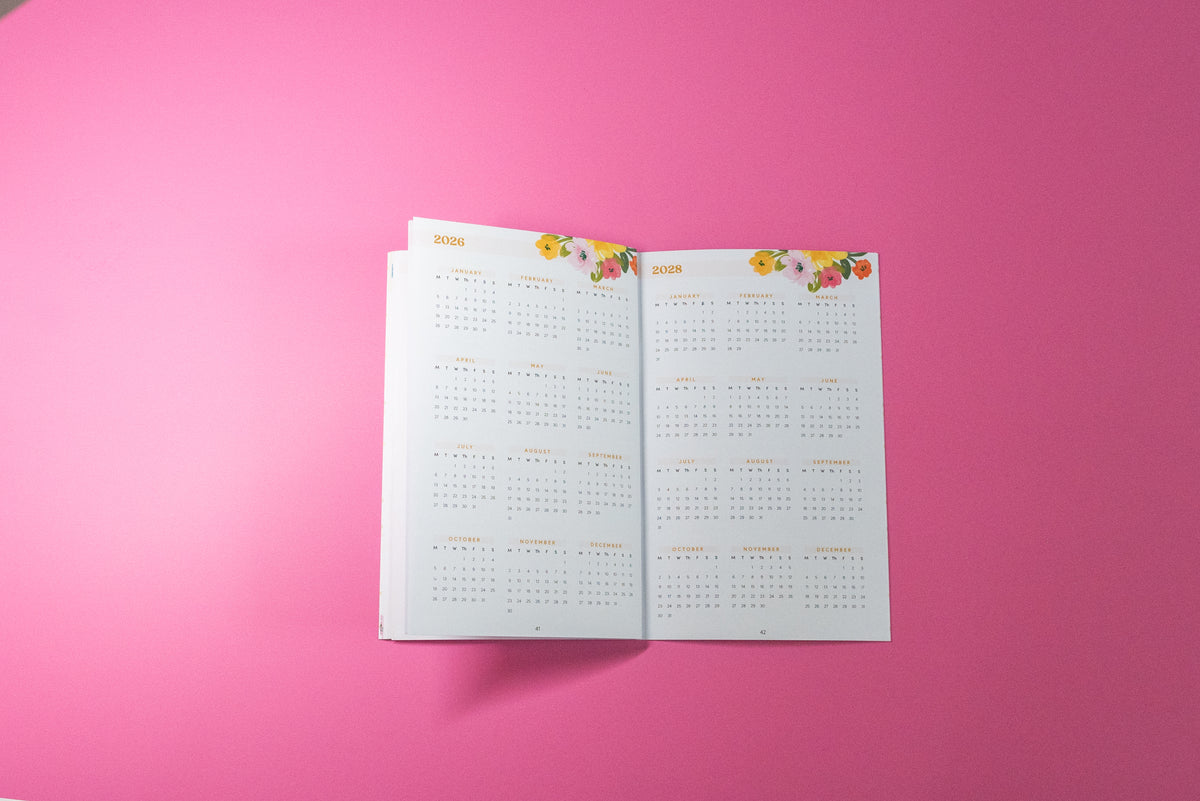 An open monthly calendar spread on a solid pink background, featuring white pages with gold text and colorful floral illustrations at the top.