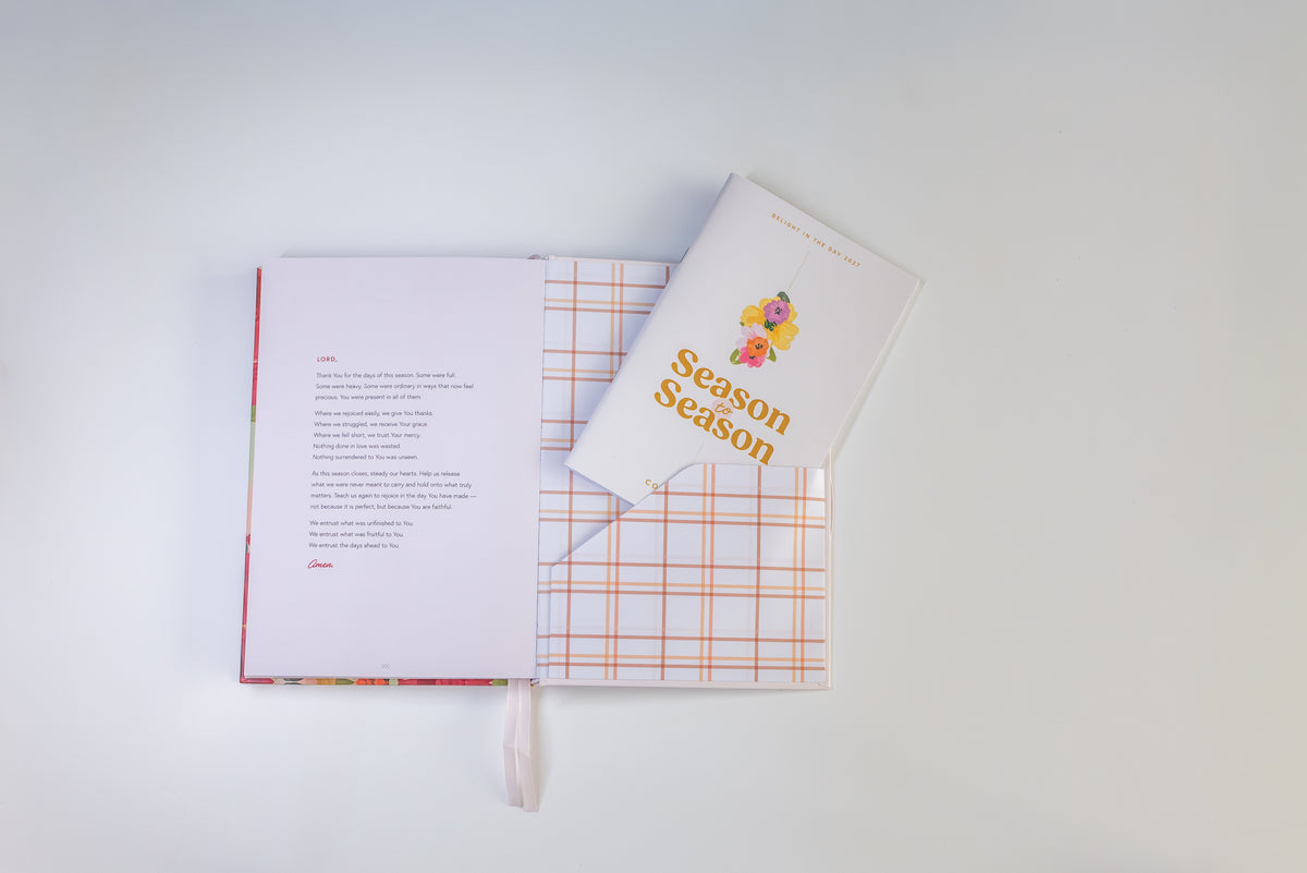 An open book with printed text on the left page and a white cover titled 'Season Season' on the right, placed on top of a white surface. The book has a white cover with orange plaid pattern and a small colorful floral illustration.