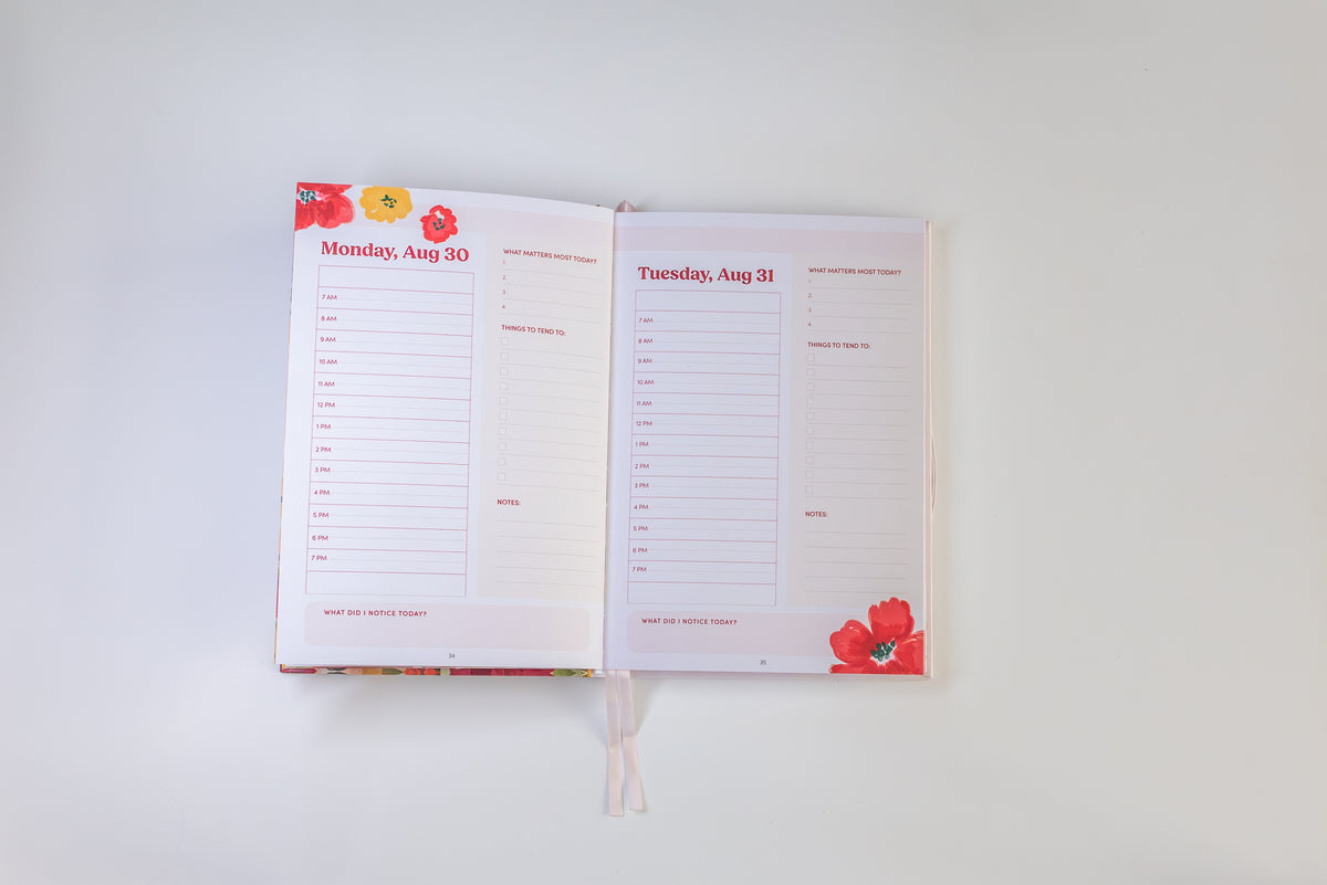 An open planner or agenda with two printed daily planning pages, photographed from above on a plain white background. The pages are mostly white with red text and small red floral illustrations, and a white ribbon bookmark hangs from the bottom center.