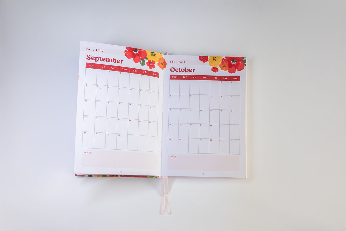 An open monthly planner showing September and October pages with red headers, floral illustrations at the top, and a white ribbon bookmark hanging from the bottom center, photographed against a plain white background.