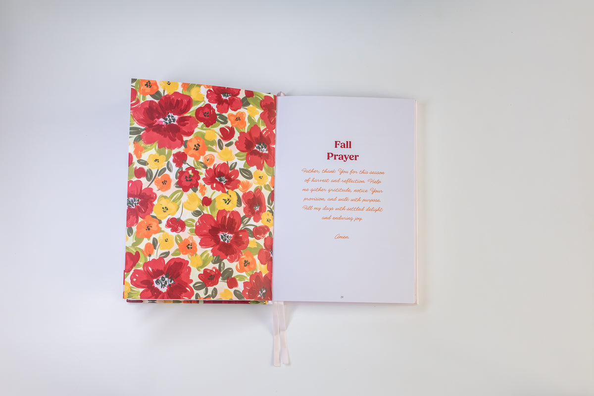 An open book or journal laid flat on a light gray background. The left page has a bright floral pattern with red, orange, yellow, and green flowers, while the right page is white with the heading 'Fall Prayer' and a short poem printed in red and orange text.