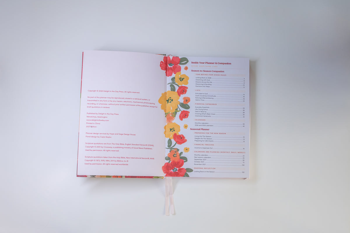 An open book or booklet laid flat on a light gray background, showing printed text pages with a vertical floral border of red, orange, and yellow flowers with green leaves along the center. A white ribbon bookmark hangs from the bottom.
