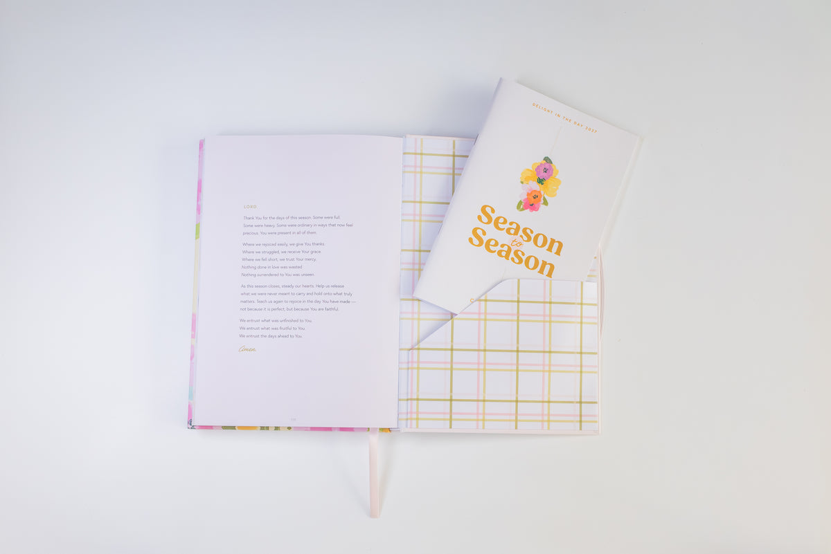 An open journal with printed pages and a matching closed copy labeled 'Season Season' lies on a white background. The journal has a pastel pink cover with a subtle floral border and a yellow-and-white plaid pattern visible on the inside pages.