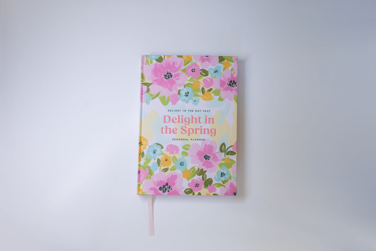 A spring-themed daily planner titled 'Delight in the Spring', featuring a colorful floral pattern in pink, yellow, and green on a white background, complete with a pink ribbon bookmark.