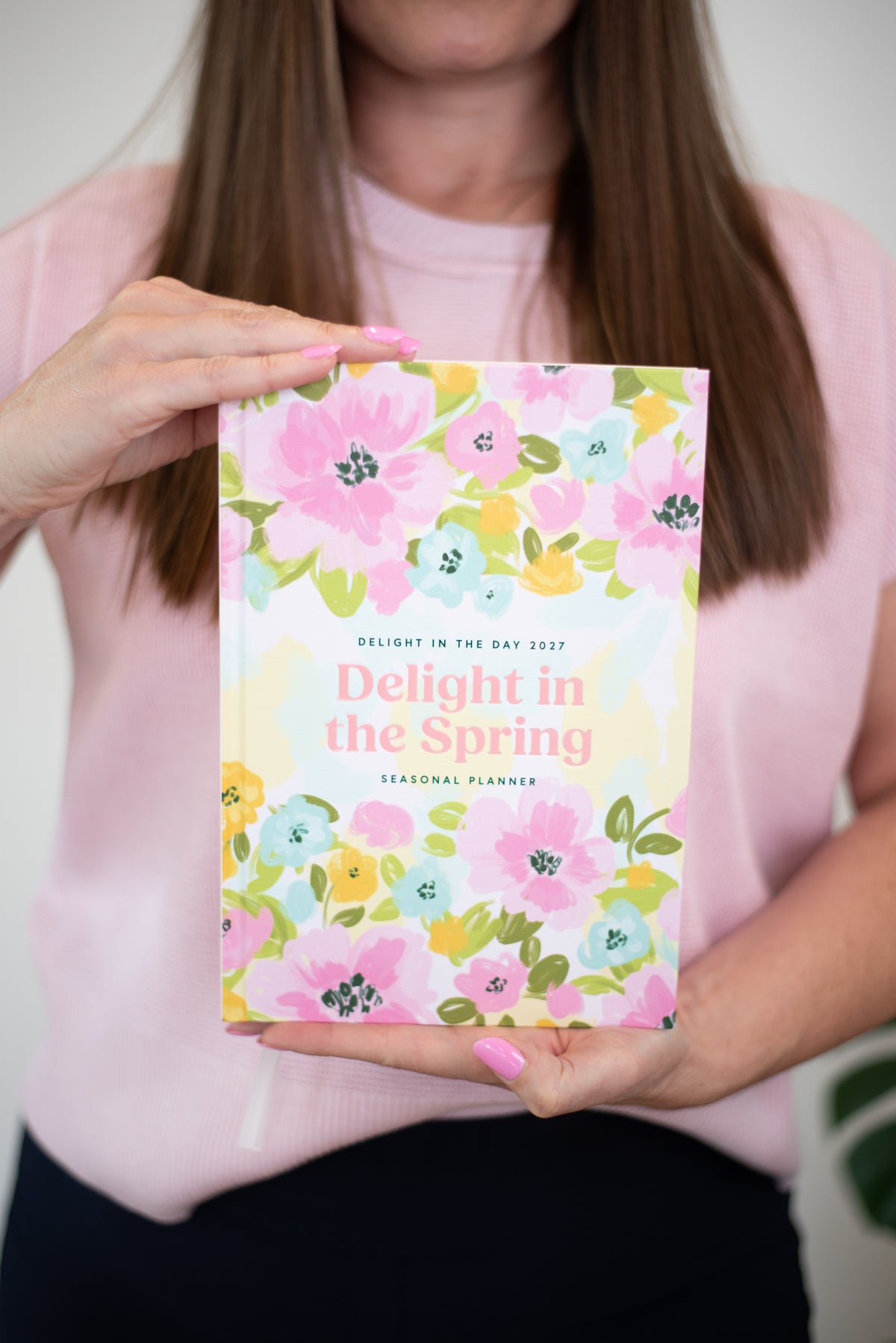 A woman in a light pink sweater holds up a floral spring seasonal planner. The planner cover features a colorful watercolor pattern of pink, yellow, and blue flowers with the title 'Delight in the Spring' in pink text.