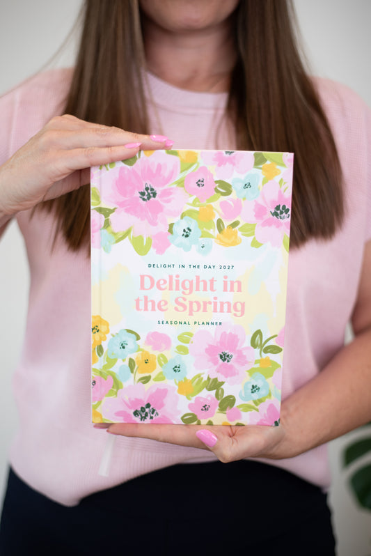 A woman in a light pink sweater holds up a floral spring seasonal planner. The planner cover features a colorful watercolor pattern of pink, yellow, and blue flowers with the title 'Delight in the Spring' in pink text.