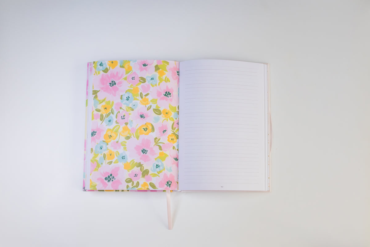 An open notebook with a pastel pink floral patterned cover on the left and lined blank pages on the right, photographed from above on a clean white background.