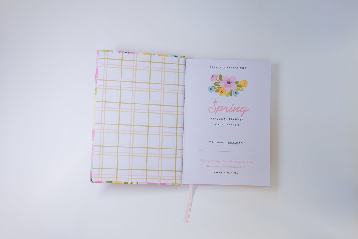 An open planner or notebook with a pastel plaid patterned cover on the left and a printed invitation page on the right featuring a floral spring-themed design, photographed on a clean white background.