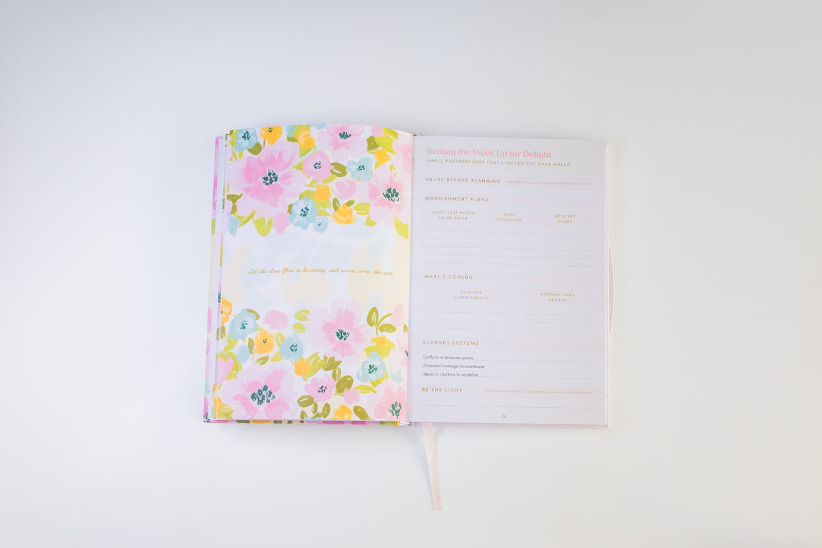 An open floral planner or journal on a clean white background, showing a pink floral-patterned cover on the left and a lined interior page on the right with gold and pink text.