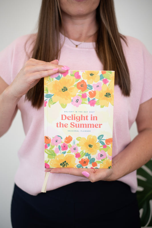 A woman in a light pink t-shirt holds up a 'Delight in the Summer' personal planner with a colorful floral cover design.