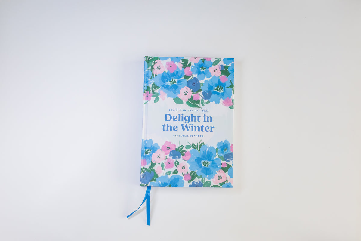 A hardcover journal titled 'Delight in the Winter', featuring a white cover decorated with blue, pink, and green watercolor flowers. A blue ribbon bookmark is tucked into the bottom edge, and the book is photographed against a clean white background.