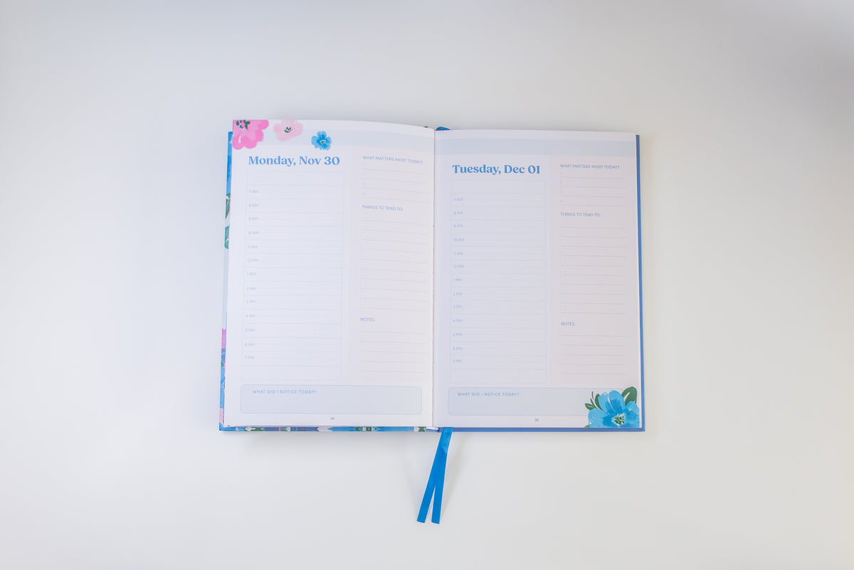 An open planner or agenda with two lined daily pages visible, decorated with floral accents in pink, blue, and green. The ribbon bookmarks extend downward, and the item is photographed from above on a plain white background.