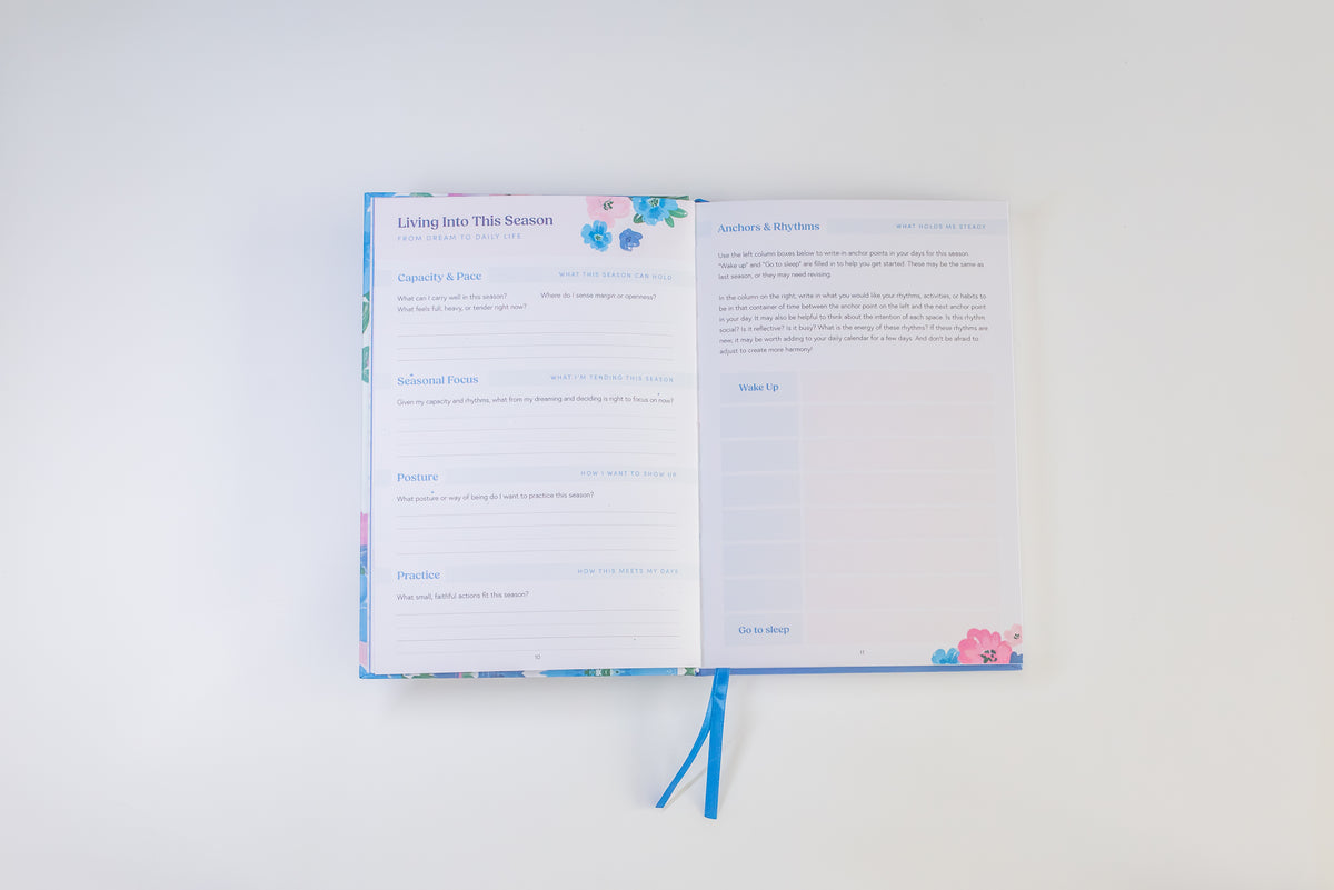 An open planner or journal with printed pages titled 'Living Into This Season' and a blue ribbon bookmark, photographed on a clean white background.