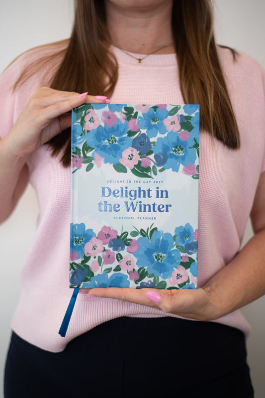 A person in a pink sweater holds a floral-bound journal titled 'Delight in the Winter' against a plain light background.