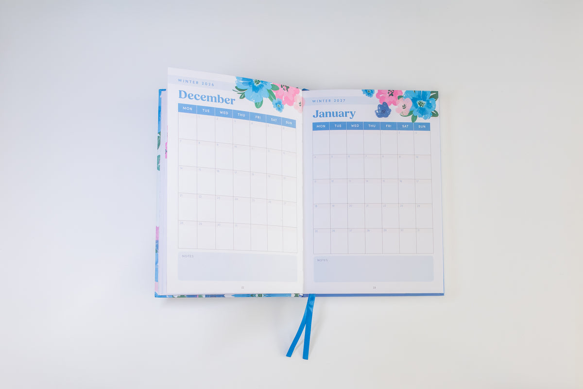 An open planner or desk calendar showing the December and January monthly grids, with floral accents at the top and ribbon bookmarks hanging below, photographed against a plain white background.