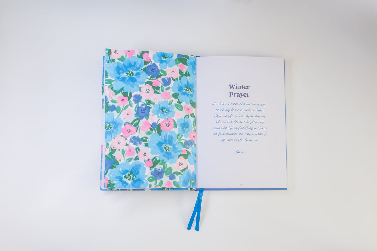 An open book with a floral-patterned cover in blue, pink, and green on one side and a printed page titled 'Winter Prayer' on the other, with blue ribbon bookmarks visible, photographed on a clean white background.