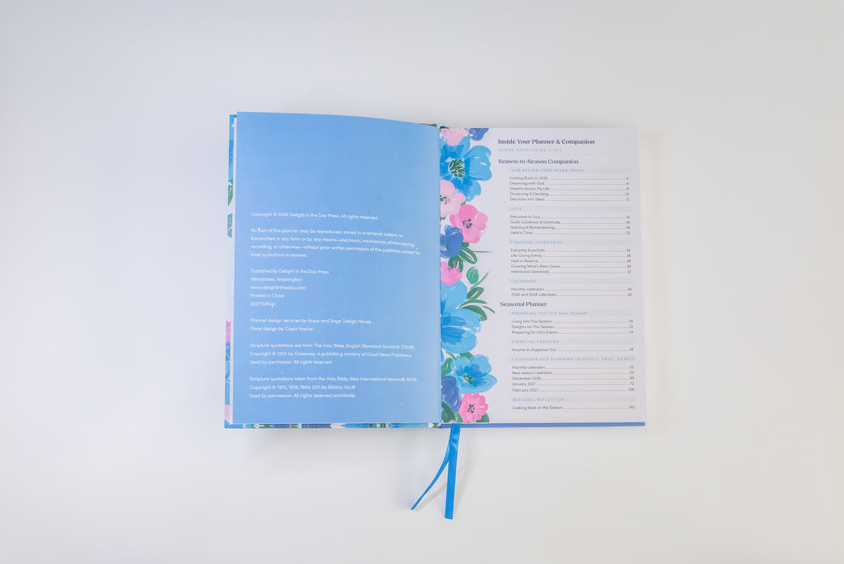 An open book or journal laid flat on a white background, showing a light blue left page with text, a floral border with pink and blue flowers, and a right page with a table layout. A blue ribbon bookmark hangs from the bottom center.