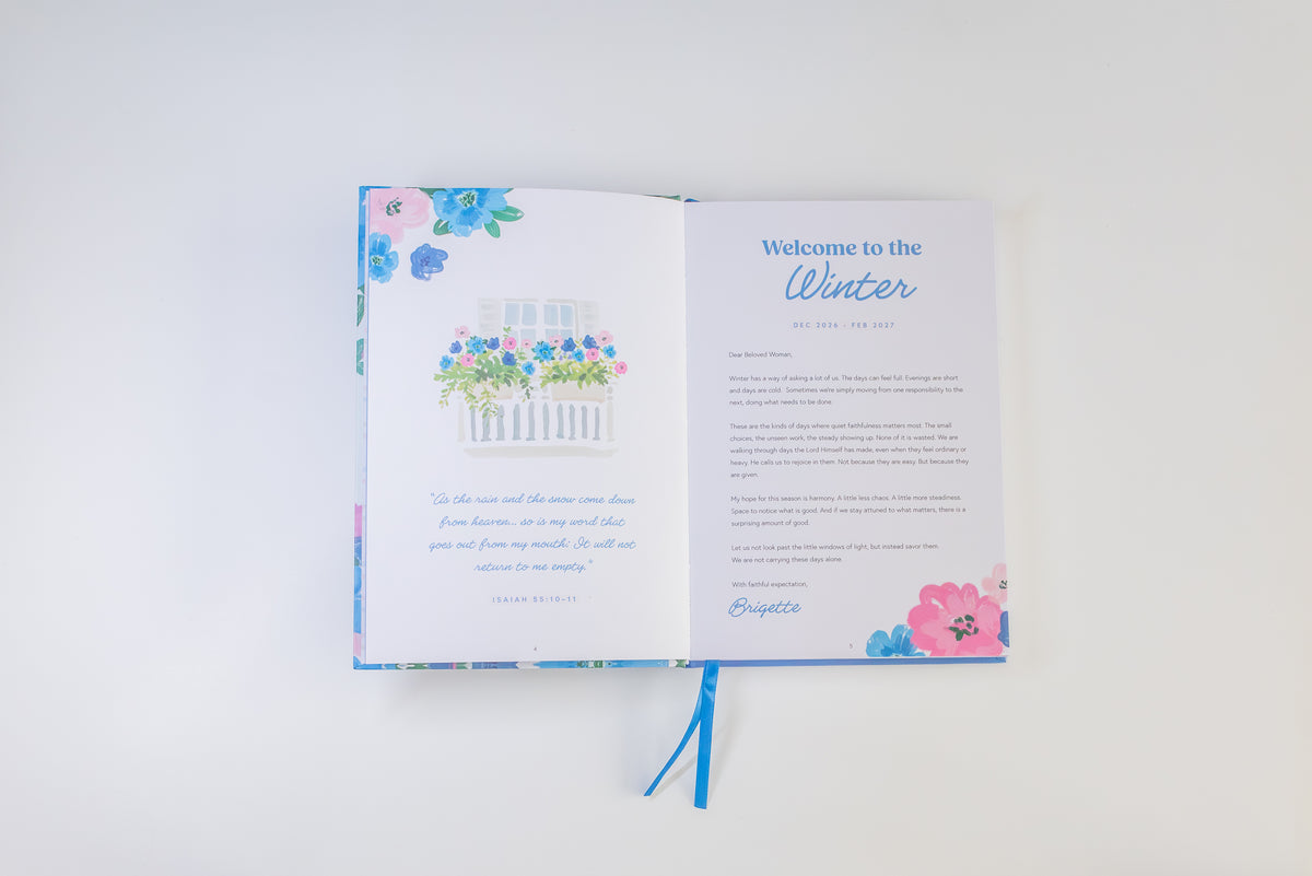 An open printed journal or gift book with floral blue and pink page designs, shown on a clean white background. The left page features a small flower illustration above a quote, and the right page has a 'Welcome to the Winter' heading with body text and a blue ribbon bookmark.