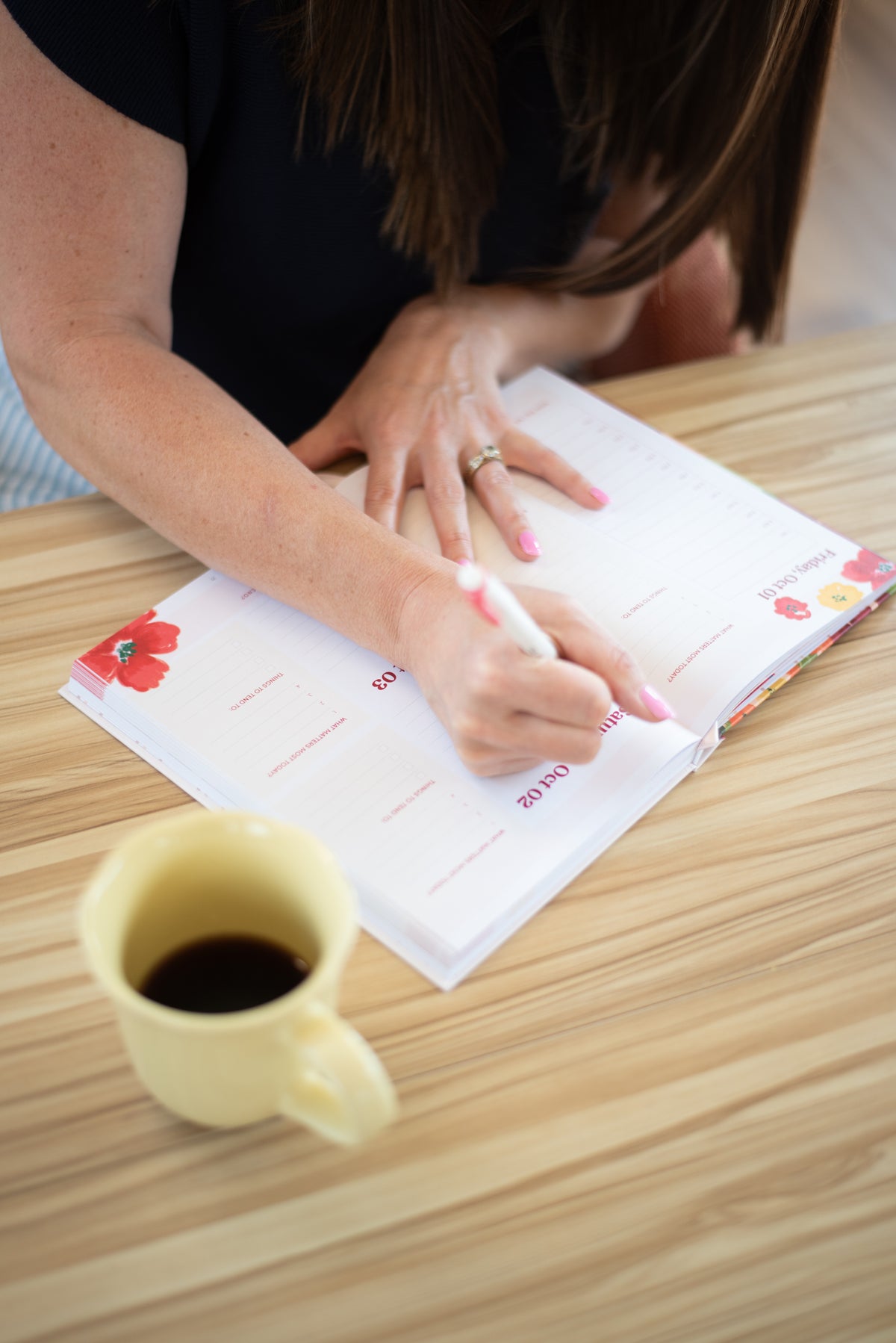 A person writing in a white planner or journal on a light wood table, with a yellow mug in the foreground and the person's arms and hands visible.