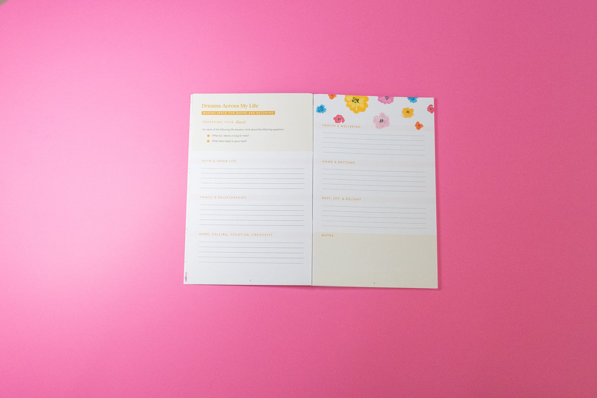 An open planner or notebook showing two interior pages with lined writing areas, cream headers, and small colorful floral stickers along the top right, photographed on a solid pink background.