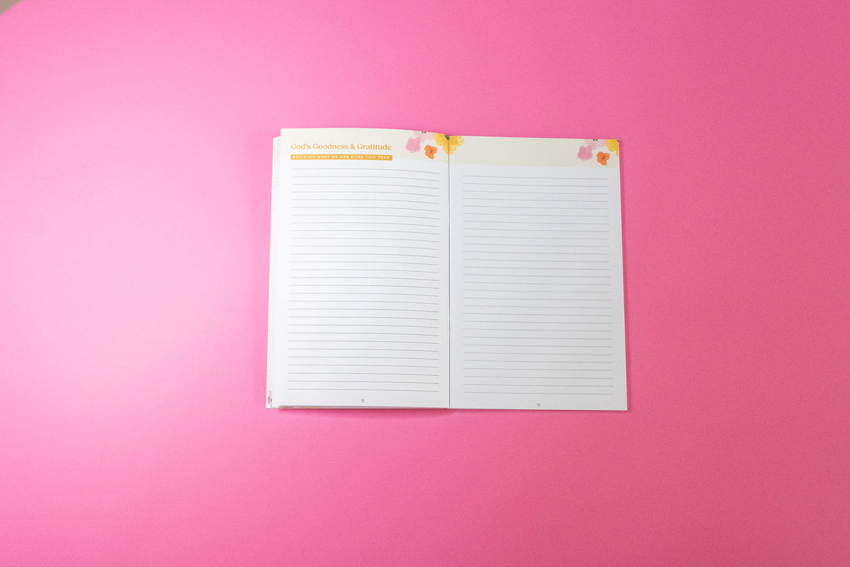 An open lined notepad with two white pages, small decorative floral corner accents, and a gold header on the left page, photographed on a solid pink background.