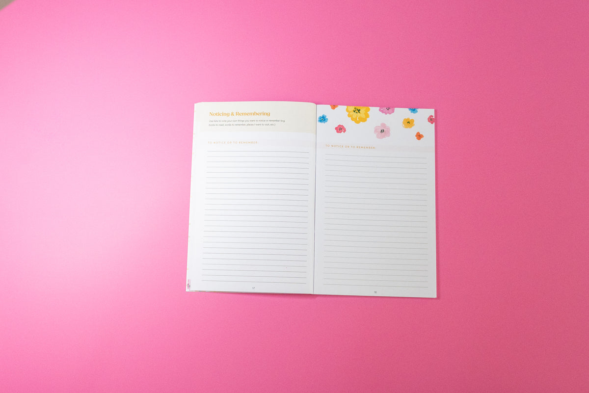 An open lined journal or notebook with two pages spread, placed on a solid pink background. The left page has a cream header, and the right page has a floral border with small colorful flowers at the top.