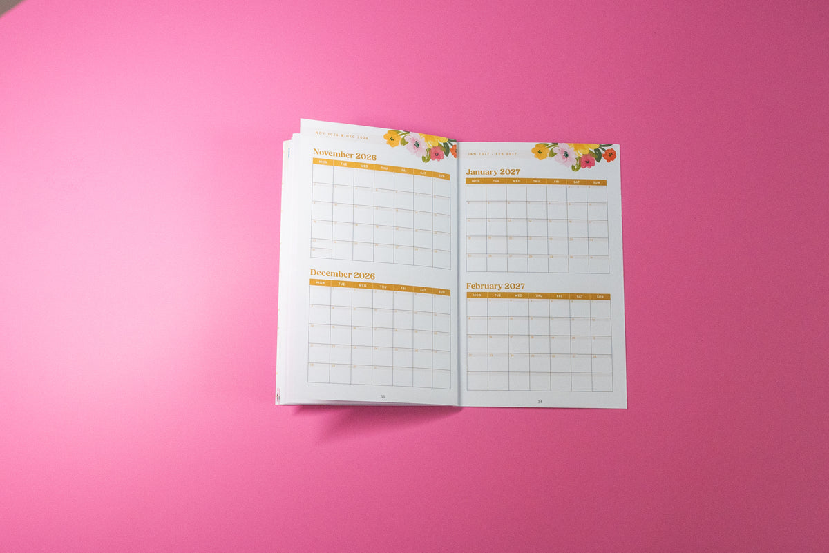 An open monthly planner showing pages for November 2026 through February 2027 with floral accents at the top, photographed on a solid pink background.
