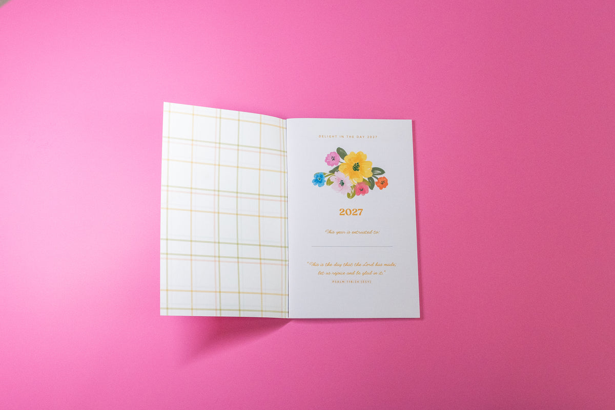 An open greeting card or invitation on a solid pink background. The left page has a white and gold grid pattern, and the right page features a colorful watercolor flower illustration above the year '2027' and a quote.